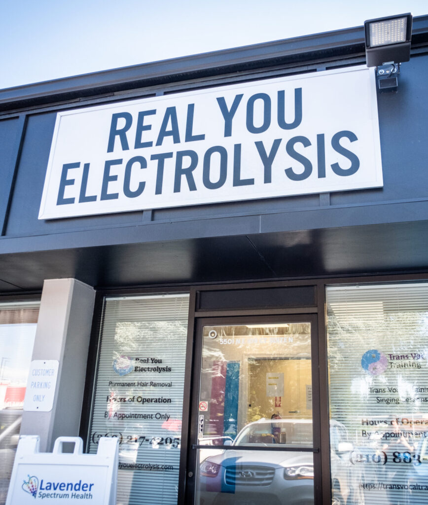 The front of the Vancouver WA location for Real You Electrolysis. There is a glass front door with a large billboard sign overhead showing the company name. The company sign above the door has a white background and large black letters. 